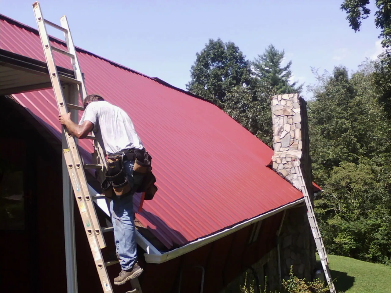 Roofer on a ladder beside a red metal roof and stone chimney — Soffit & Fascia Repair by Roofing Brothers in Callicoon Center